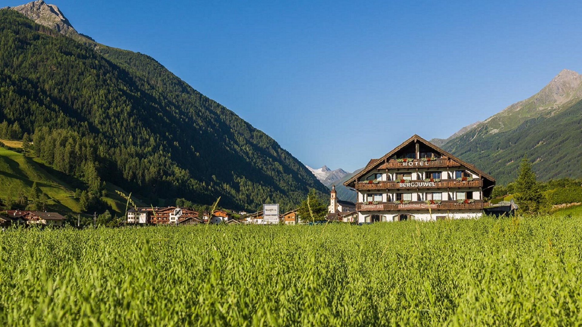 Willkommen im Hotel in Neustift im Stubaital! Hotel in grüner Wiese mit Bergen und klarem blauen Himmel im Hintergrund